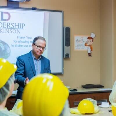 A man in a suit gestures while presenting at a podium in front of a projected slide reading "Leadership Dickinson" with yellow hard hats displayed on a table in the foreground.